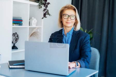 Portrait of smiling 50s stylish, confident mature businesswoman, middle aged company ceo director, experienced senior female professional, business coach working on laptop in office. Female leader.