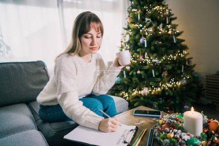 Photo for Young Woman with cup is hand writing New years goals on note paper in new year day. New year resolutions list. Goals, resolutions, plans, action, checklist concept. New Year template, copy space. - Royalty Free Image