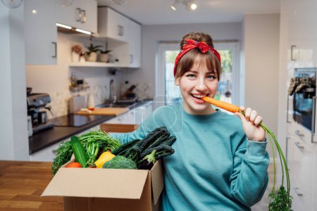 Smiling woman eating fresh carrot from her healthy food delivery box. Box with vegetables and phone with Veganuary app on the kitchen table. Start of a healthy life concept. Online home food delivery