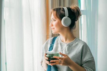 Young woman listen music, enjoy cup of coffee or tea and looks out window. Calm female spend free time at home enjoy favourite song with wireless headphones. Pastime weekend relax, no stress concept