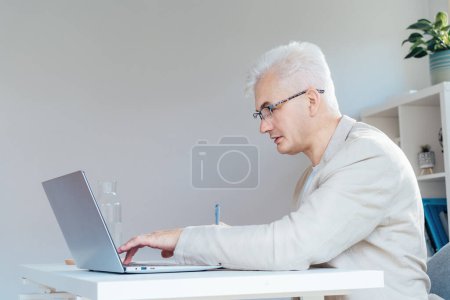 Foto de El hombre de mediana edad de pelo gris concentrado toma notas mientras trabaja en la computadora portátil en el lugar de trabajo en la oficina en el hogar. Confiado, experimentado profesional masculino senior. Empresario de pequeñas empresas administrar negocios - Imagen libre de derechos