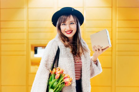 Photo for Portrait of smiling fashion woman with box near modern postal automatic mail terminal with self service device for pickup or refund an order. Electronic locker for storing parcels. Selective focus. - Royalty Free Image