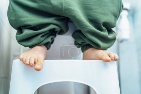 Téléchargez les photos : Gros plan mâle adulte assis sur des toilettes blanches avec son pantalon baissé et ses pieds sont debout sur le pied de toilette Tabouret pour un processus facile de selles, allant aux toilettes pour faire caca. Concentration sélective - en image libre de droit