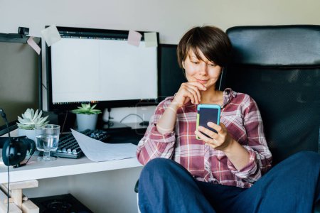 Photo for Smiling woman looking at the phone screen while sitting on her home office or office working place. Take a break. Reading the good news. Remote work, social networks, blogging. Telecommuting concept - Royalty Free Image