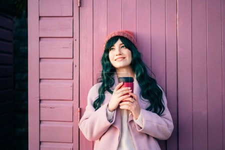 Relajada joven y elegante mujer hipster sonriente con pelo de color, abrigo rosa, sombrero que sostiene taza de café reutilizable apoyado en la cabaña de la playa y disfrutar del momento. Placeres simples y realización personal