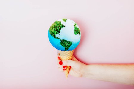 Téléchargez les photos : Femme main tenant un cône de gaufre avec la planète Terre modèle comme dessert à la crème glacée sur fond rose. Concept de réchauffement, trou d'ozone, fonte, pollution ou changement climatique. Sauvez la planète, concept de jour de la Terre - en image libre de droit