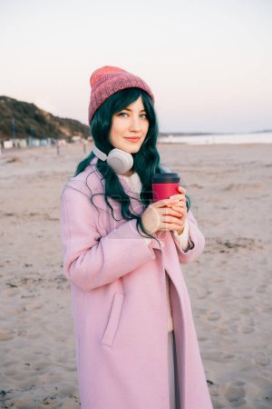 Elegante joven mujer hipster sonriente con el pelo de color con auriculares inalámbricos, abrigo rosa, sombrero, caminando en la playa con taza de café reutilizable. Estilo de vida solitario, tendencia de estilo de moda estacional. Vertical.