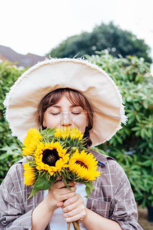 Dreaming young female farmer woman with closed eyes holding and sniffing a sunflowers bouquet on the green garden background. Enjoy the moment. Rural, Cottage core lifestyle. Vertical card.