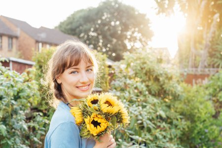 Portrait of smiling tender young woman holding fresh sunflowers bouquet and enjoying carefree moment in sunset light. Rural farm life, Cottage core lifestyle. Simple pleasures. Selective focus
