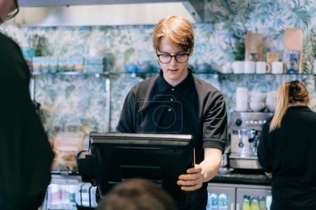 Young waiter serving customer at cash point in cafe. Man working with POS terminal. Cashier, barista checking for payment receipt. Hospitality, server and preparing a slip at the till in coffee shop