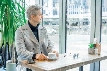 Portrait of thoughtful handsome young stylish man in checked coat relaxing with cup of coffee in modern cafe shop . Cold season elegant fashion stylish outfit trend. Retro, Old money aesthetic
