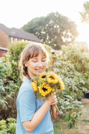 Portrait of smiling tender young woman holding fresh sunflowers bouquet and enjoying carefree moment in sunset light. Rural farm life, Cottage core lifestyle. Selective focus. Vertical card.