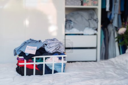 Auf dem Bett steht ein Korb mit ausgewählten Kleidungsstücken, die für den Charity-Laden gespendet werden können. Kleidung entschlüsseln, sortieren und aufräumen. Wiederverwendung aus zweiter Hand. Bewusster Verbraucher, Nachhaltigkeit. Selektiver Fokus