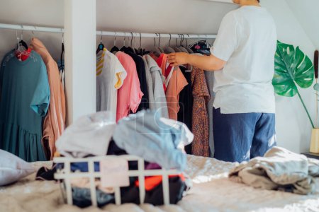 Frau sucht Kleidung aus ihrem Kleiderschrank aus, die sie für einen Charity-Laden spenden möchte. Deklarieren, Kleider sortieren und aufräumen. Wiederverwendung, Second-Hand-Konzept. Bewusster Verbraucher, nachhaltiger Lebensstil