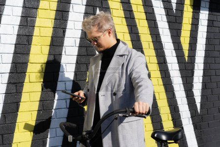 Stylish male in coat and glasses using phone while walking with retro bicycle by painted urban wall in sunny day. Neutral carbon footprint transportation. Ecofriendly mobility sustainable transport.