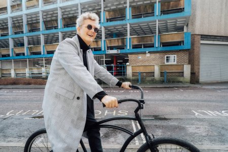 Stylish male in checked coat and sunglasses riding on his retro bicycle on street with parking buildings. Neutral carbon footprint transportation. Green eco friendly mobility sustainable transport