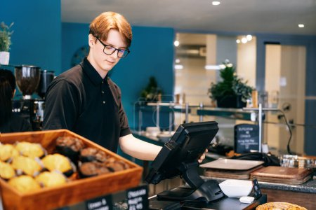 Young waiter serving customer at cash point in cafe. Man working with POS terminal. Student working part-time. Problems of unemployment and Job opportunities for youth. Small business vacancies.