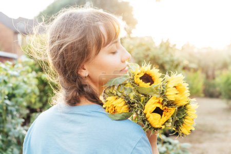 Dreaming young female farmer woman with closed eyes holding and sniffing a sunflowers bouquet on the green garden background in sunset light. Enjoy the moment. Rural, Cottage core lifestyle.