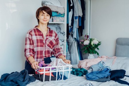 Photo for Smiling Woman with basket of selected clothes from her wardrobe for donating to Charity. Decluttering, Sorting clothes And Cleaning Up. Reuse, second-hand concept. Conscious consumer, sustainability - Royalty Free Image