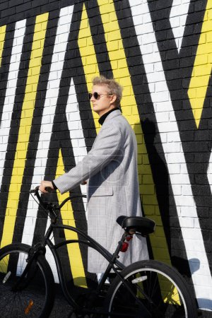 Stylish male in checked coat and sunglasses goes with his retro bicycle by painted urban wall in sunny day. Neutral carbon footprint transportation. Green eco friendly mobility sustainable transport.