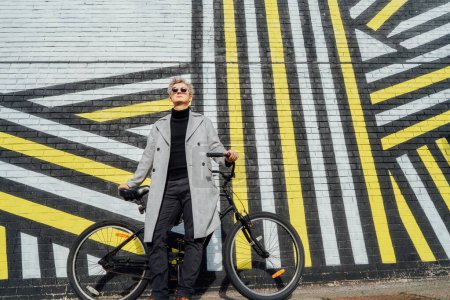 Foto de Elegante hombre de abrigo a cuadros y gafas de sol de pie con bicicleta retro por pared urbana pintada en un día soleado. Transporte de huella de carbono neutro. Movilidad ecológica verde transporte sostenible. - Imagen libre de derechos