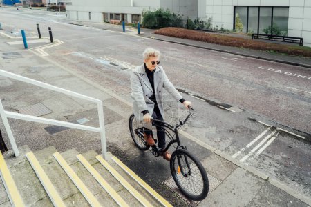 Stylish male in checked coat and sunglasses riding on his retro bicycle on the street with modern buildings. Neutral carbon footprint transportation. Green eco friendly mobility sustainable transport.