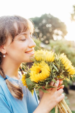 Dreaming young female farmer woman with closed eyes holding and sniffing a sunflowers bouquet on the green garden background in sunset light. Enjoy the moment. Rural, Cottage core lifestyle. Vertical.