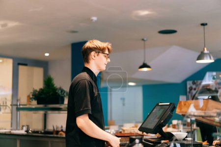 Young waiter serving customer at cash point in cafe. Man working with POS terminal. Cashier, barista checking for payment receipt. Hospitality, server and preparing a slip at the till in coffee shop.
