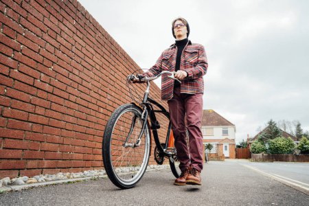 Junger, stylischer Mann fährt mit seinem Retro-Fahrrad an einer Ziegelmauer der Vorstadt zwischen Häusern vorbei. Student auf dem Weg zur Hochschule mit dem Fahrrad. Umweltfreundlicher Transport für die CO2-Bilanz. Nachhaltiger Lebensstil.