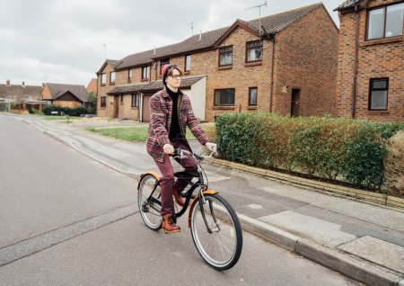 Young stylish hipster man riding on city Bicycle on the road of suburbs area among houses. Student on way to college on bike. Eco friendly transportation for carbon footprint. Sustainable lifestyle.