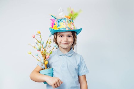 Oeufs de Pâques chasse tradition. Un garçon souriant dans un chapeau de Pâques tient un arbre de Pâques en pot et un oeuf en chocolat sur fond blanc. Décoration de Pâques, célébration familiale, traditions chrétiennes