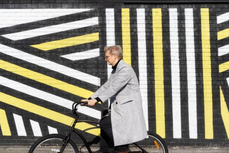 Foto de Elegante hombre de abrigo a cuadros y gafas de sol montado en su bicicleta retro por la pared urbana pintada en un día soleado. Transporte de huella de carbono neutro. Movilidad ecológica verde transporte sostenible. - Imagen libre de derechos
