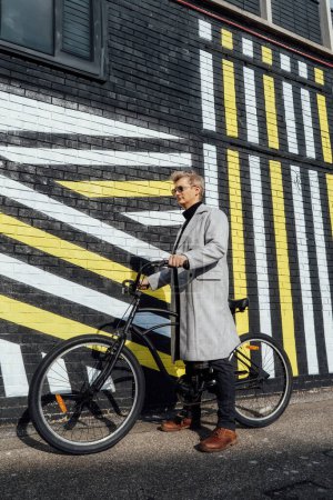 Stylish male in checked coat and sunglasses standing with retro bicycle by painted urban wall in sunny day. Neutral carbon footprint transportation. Green eco friendly mobility sustainable transport.