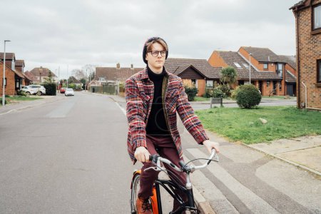 Young stylish hipster man riding on city Bicycle on the road of suburbs area among houses. Student on way to college on bike. Eco friendly transportation for carbon footprint. Sustainable lifestyle.