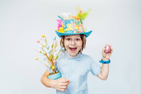 Easter eggs hunt tradition. Emotionally shouting boy kid in Easter hat holds easter tree in pot and shaking chocolate egg in other hand on the white background. Easter decoration and traditions