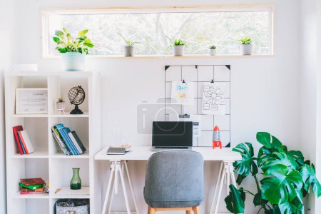Modern cozy light workplace - white desk with laptop black screen, grid mood board with pinned notes, shelves with docs and green monstera plant at work space in home office room interior.