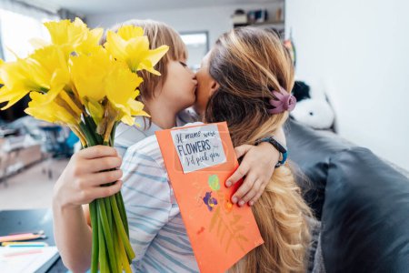 Glücklicher Muttertag. Nahaufnahme Porträt eines lächelnden Jungen, der seine Mutter umarmt, nachdem er ihr gratuliert und ihr Blumen und handgemachte Postkarten überreicht. Familienurlaub und Zweisamkeit