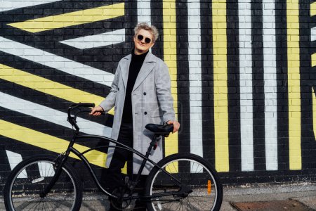 Stilvoller Mann in kariertem Mantel und Sonnenbrille steht mit Retro-Fahrrad an der bemalten Stadtmauer bei sonnigem Tag. Neutraler Transport von CO2-Fußabdrücken. Grüne umweltfreundliche Mobilität nachhaltiger Verkehr.