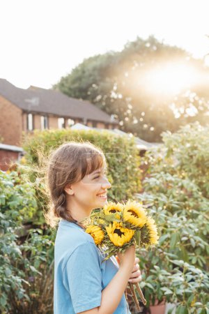 Portrait d'une jeune femme souriante tenant un bouquet de tournesols frais et profitant d'un moment d'insouciance au coucher du soleil. Ferme rurale, Cottage style de vie de base. Concentration sélective. Carte verticale.