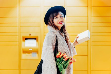 Photo for Portrait of smiling fashion woman with box near modern postal automatic mail terminal with self service device for pickup or refund an order. Electronic locker for storing parcels. Selective focus - Royalty Free Image