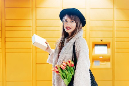 Portrait of smiling fashion woman with box near modern postal automatic mail terminal with self service device for pickup or refund an order. Electronic locker for storing parcels. Selective focus