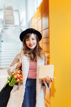 Photo for Portrait of smiling fashion woman with box near modern postal automatic mail terminal with self service device for pickup or refund an order. Electronic locker for storing parcels. vertical card. - Royalty Free Image