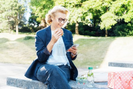 Middle-aged business woman eating sandwich burger with salad and using phone sitting on bench in the park while having lunch break. Remote work,business, freelance,blogging,social media concept