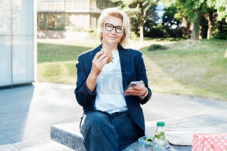 Middle-aged business woman eating sandwich burger with salad and using phone sitting on bench in the park while having lunch break. Remote work,business, freelance,blogging,social media concept.