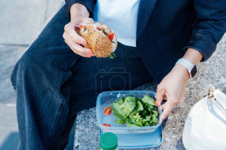 Téléchargez les photos : Vue de dessus gros plan femme d'affaires manger un sandwich et salade fraîche sur la pause déjeuner assis sur le banc au parc. Boîte à lunch régime équilibré pour la perte de poids. Habitudes alimentaires saines et bien-être. Concentration sélective - en image libre de droit
