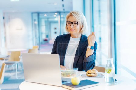 Middle-aged business woman working on laptop and having healthy lunch at working place, office cafe. Alternative office lifestyle female working in video call conference during lunch time. Modern job.