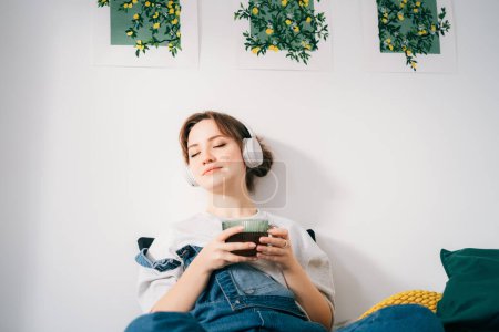 Woman with closed eyes listen music and enjoy cup of coffee or tea. Calm female spend free time at home enjoy favorite song with wireless modern headphones. Pastime weekend relax, no stress concept.