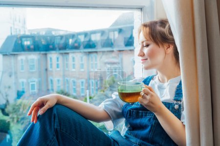 Time To Relax. Young woman looks out the window overlooking the city, sits on the windowsill at cozy home holds cup of hot tea drink. Happy calm female taking break for mental health wellbeing