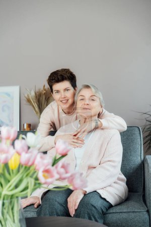 Senior 70s mother and adult neutral gender daughter hugging. Happy family enjoying weekend together in living room. Elderly woman spending quality time with daughter who visits her in retiring house