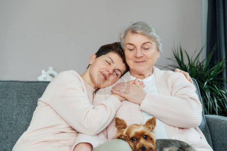 Smiling neutral gender woman and senior mother hugging, elderly mother and adult daughter enjoying tender moment, hugging, holding hands, happy family spending leisure time at home together.